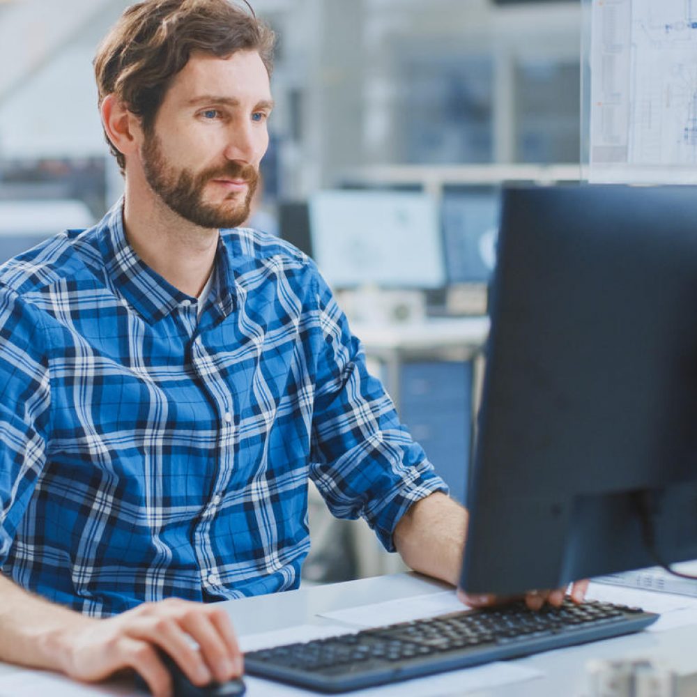 In the Industrial Engineering Facility: Portrait of the Handsome Smart Male Engineer Working on a Desktop Computer. In the Background Specialists and Technicians Working with Drafts and Drawings