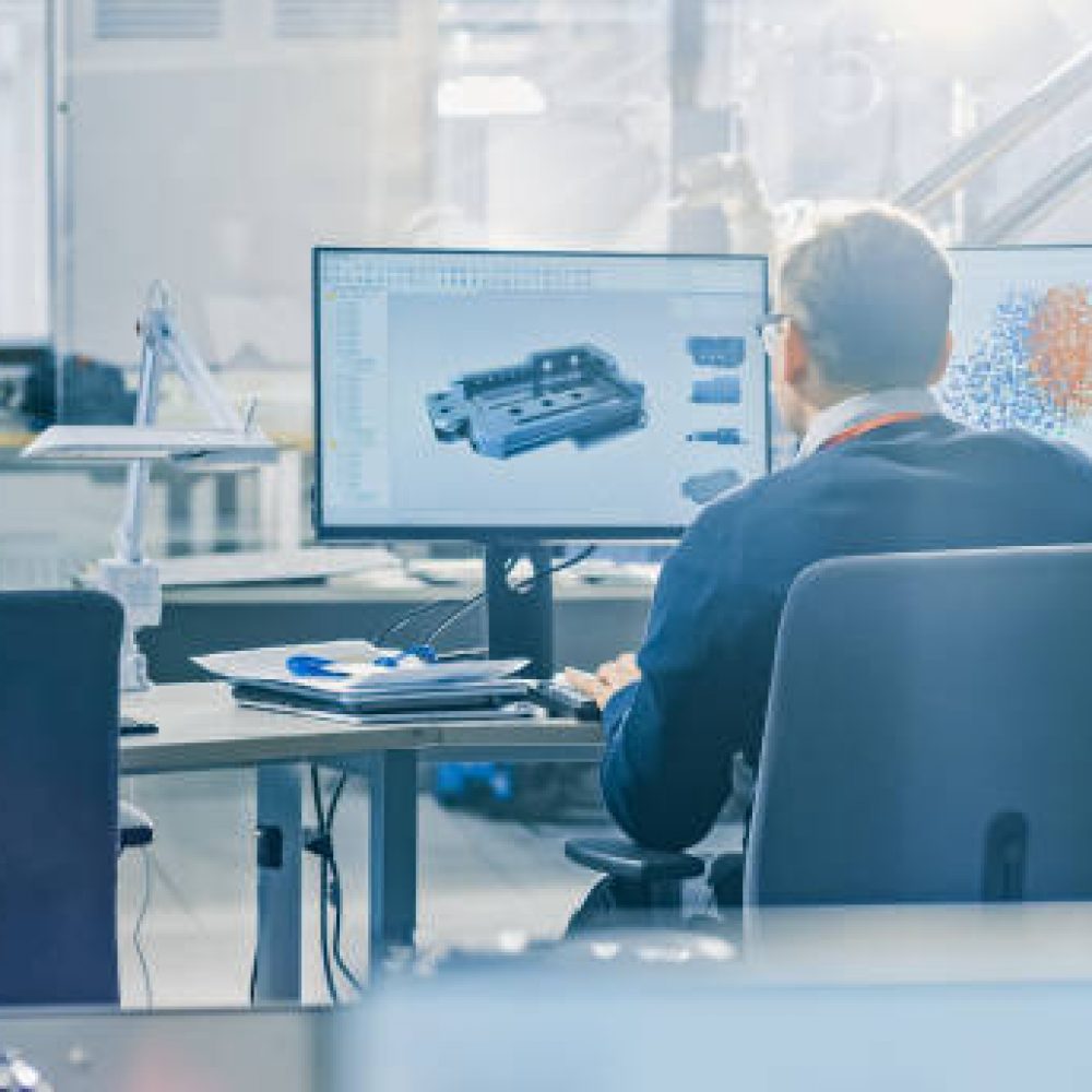 Back View of Industrial Engineer Working on Desktop Computer in Bright Office. Screens Show IDE / CAD Software, Implementation of Machine Learning, Neural Networking and Cloud Computing