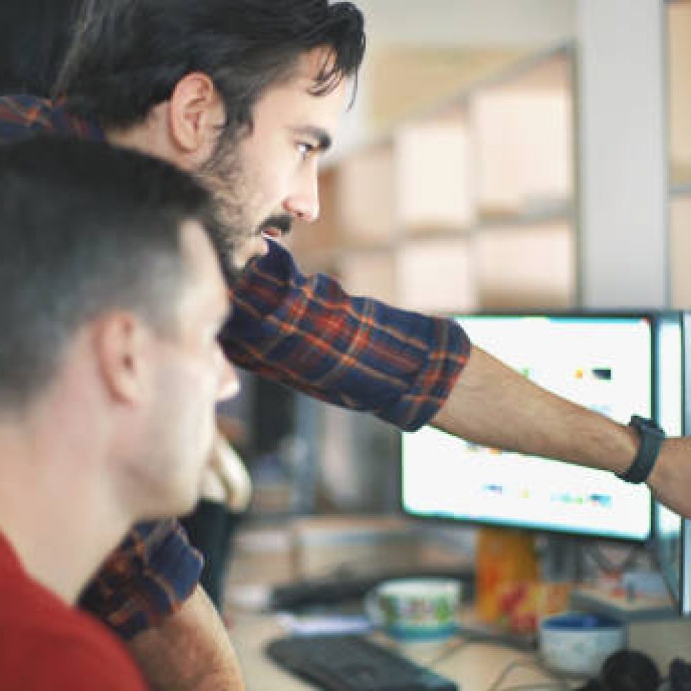 Closeup side view of group of mid 30's web designers working on a project. They are gathered around desktop computer and discussing a project. One of the guys is explaining something and pointing at computer screen. There are two men in focus and some blurry coworkers in background.